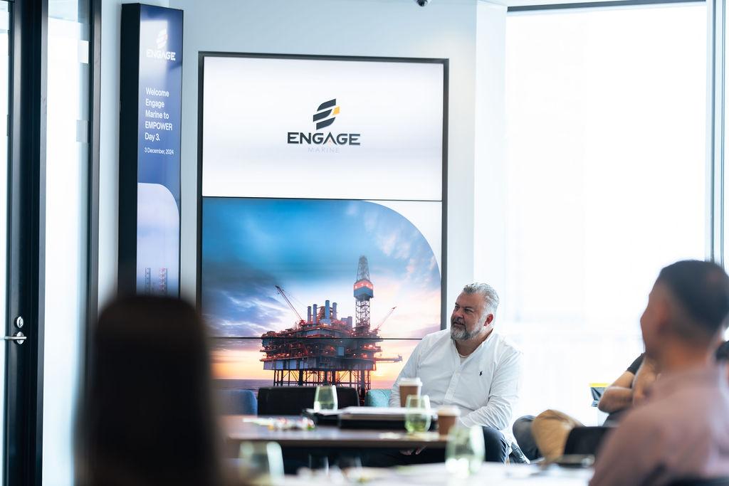 Conference setting featuring a speaker seated at a table, with the Engage Marine logo and an image of an offshore oil rig displayed prominently on a screen in the background. The room is bright and filled with natural light, with participants seated and engaged.