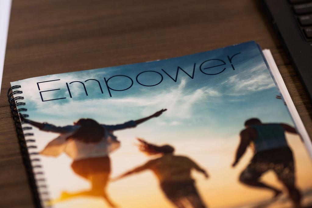 Close-up of a spiral-bound booklet on a wooden desk, featuring the word 'Empower' on the cover. The cover image shows silhouettes of people joyfully running under a bright, cloudy sky, evoking energy and optimism.