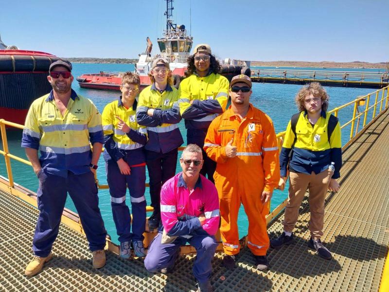 Engage Marine staff and a group of boys from the Clontarf Foundation pose together on a dock in Dampier, with the ocean and tugboats in the background, during a tour of Engage Marine's workshop and vessels.