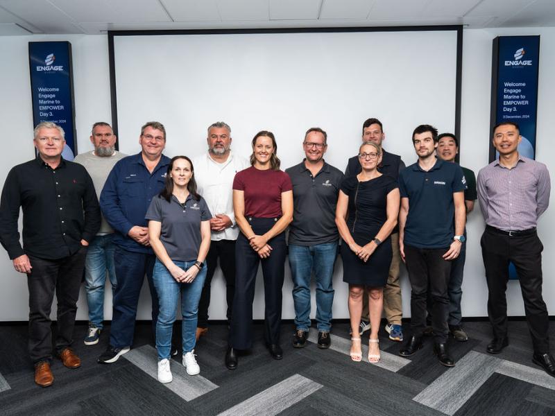 Group photo of a diverse team standing together in a conference room. The background features two vertical banners with the text 'Engage Marine' and 'Welcome Engage Marine to EMPOWER Day 3, 3 December 2024.