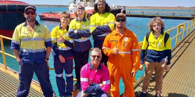 Engage Marine staff and a group of boys from the Clontarf Foundation pose together on a dock in Dampier, with the ocean and tugboats in the background, during a tour of Engage Marine's workshop and vessels.