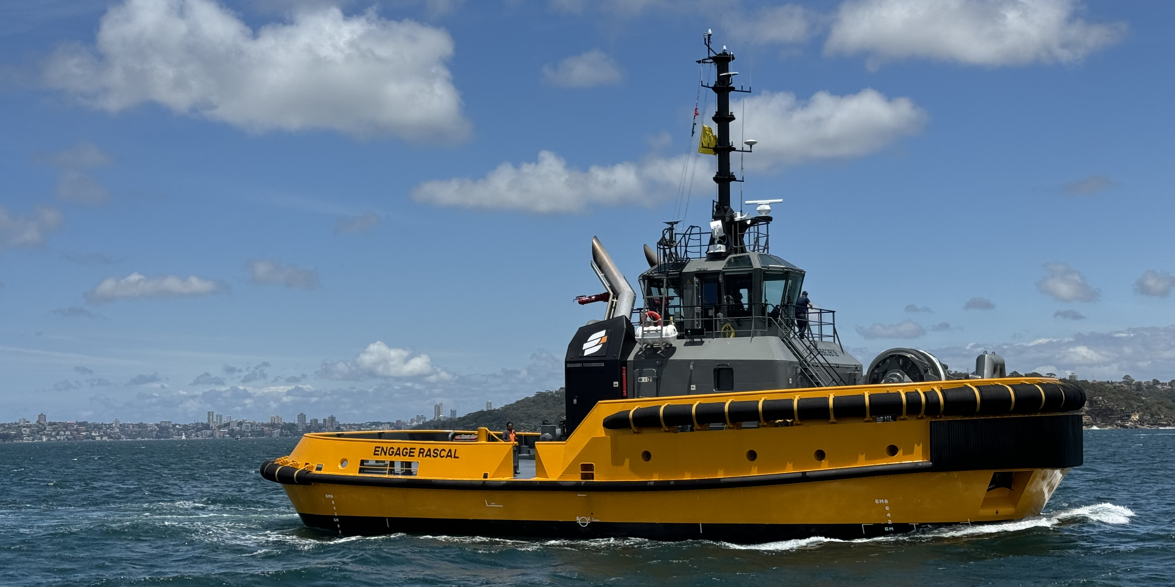 Engage Rascal, a DAMEN AD TUG 2813, navigating through calm waters under a partly cloudy sky with a distant city skyline visible in the background.