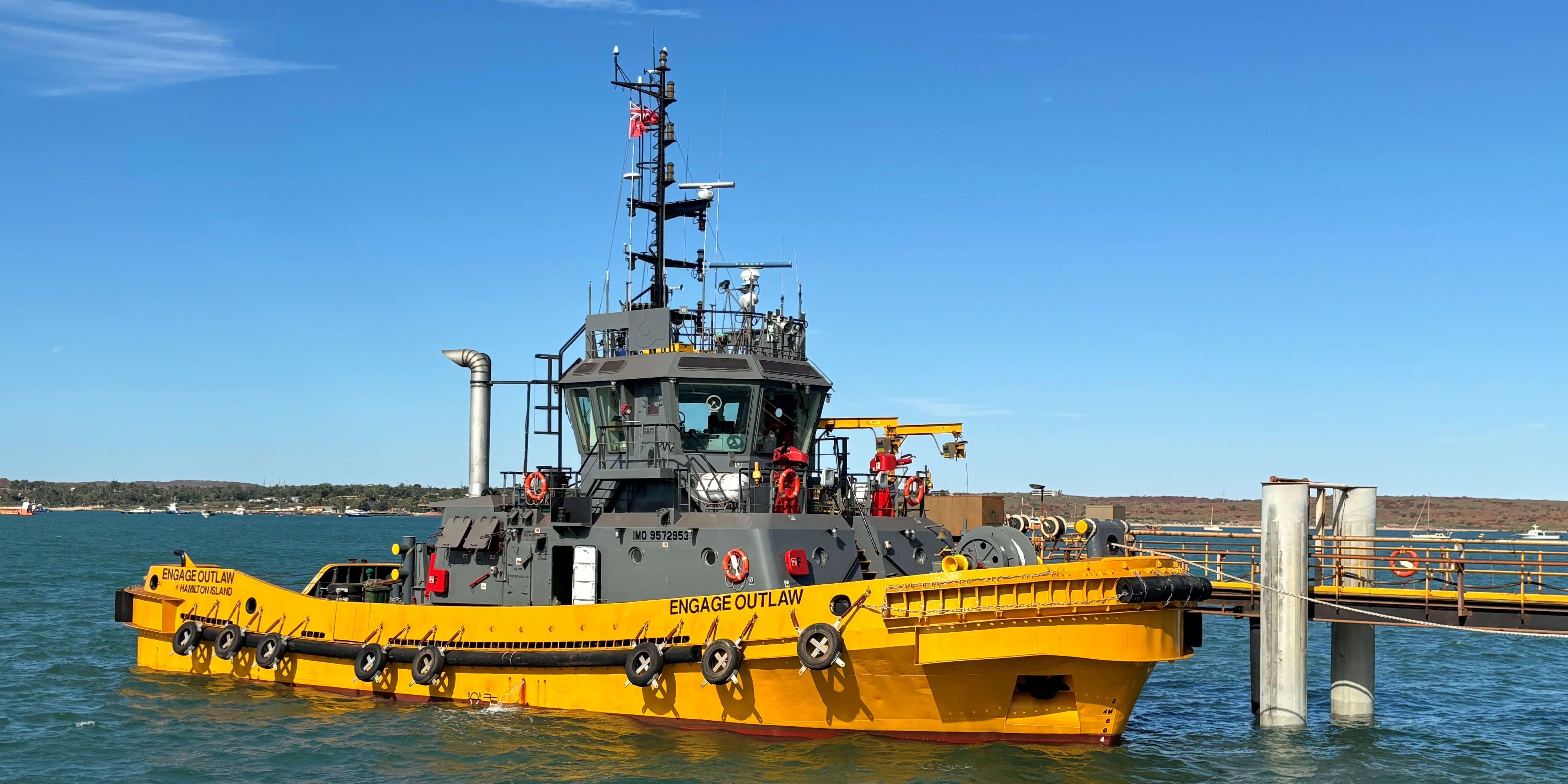 Engage Outlaw tugboat docked at port, with a clear blue sky and calm waters in the background. The bright yellow and grey vessel is seen alongside a pier, ready for towing operations.